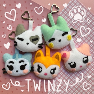 May include: Five felt animal keychains with different colors and designs. The keychains are a white cat with gray ears, a green cat, a white cat with pink ears, a yellow fox, and a blue cat. The keychains are on a pink and white background with hearts and the word "Twinzy".