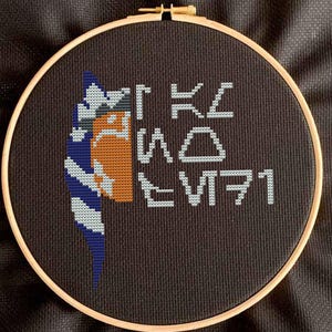 May include: A cross-stitch embroidery hoop featuring a design with a character's face in orange, white, and blue, and text in white. The embroidery is set against a dark brown fabric background, framed by a wooden hoop.