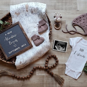 May include: A white onesie with the text "BABY Lastname" on it, a letter board with the text "A NEW Adventure Begins SEPTEMBER 20XX", a brown wooden beaded garland, a brown wicker basket with a white and gray blanket with a woodland animal print, a brown knitted baby hat, a black and white ultrasound image, and a brown wooden deer rattle.