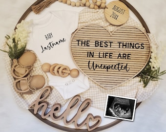 May include: A wooden tray with a white onesie, a wooden heart with the text "The best things in life are unexpected", a wooden "hello" sign, a pair of brown baby shoes, a wooden rattle, a small white blanket, a brown and white checkered blanket, a wooden bead garland, a wooden circle with the text "August 20XX", and a black and white ultrasound image.