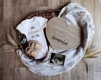 Op de afbeelding: Een witte romper met de tekst "BABY Lastname JULY, 20XX" in gouden letters. Naast de romper is een hartvormig letterbord met de tekst "THE BEST THINGS IN LIFE ARE UNEXPECTED". Een kleine blauwe speen, twee paar bruine babyschoentjes en een zwart-witte echografie zijn ook op de foto te zien.
