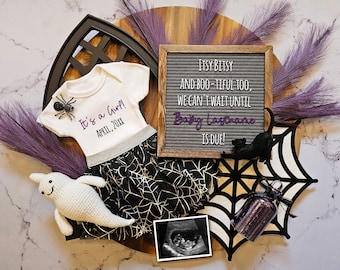 Op de afbeelding: Een Halloween-thema baby aankondiging met een witte romper met de tekst "It's a Girl! April, 20XX", een zwart-witte spinnenweb rok, een wit spook knuffeldier, een zwarte spin, een letterbord met de tekst "Itsy Bitsy and Boo-tiful too, We can't wait until Baby Lastname is due!" en een zwart kattenbeeldje.