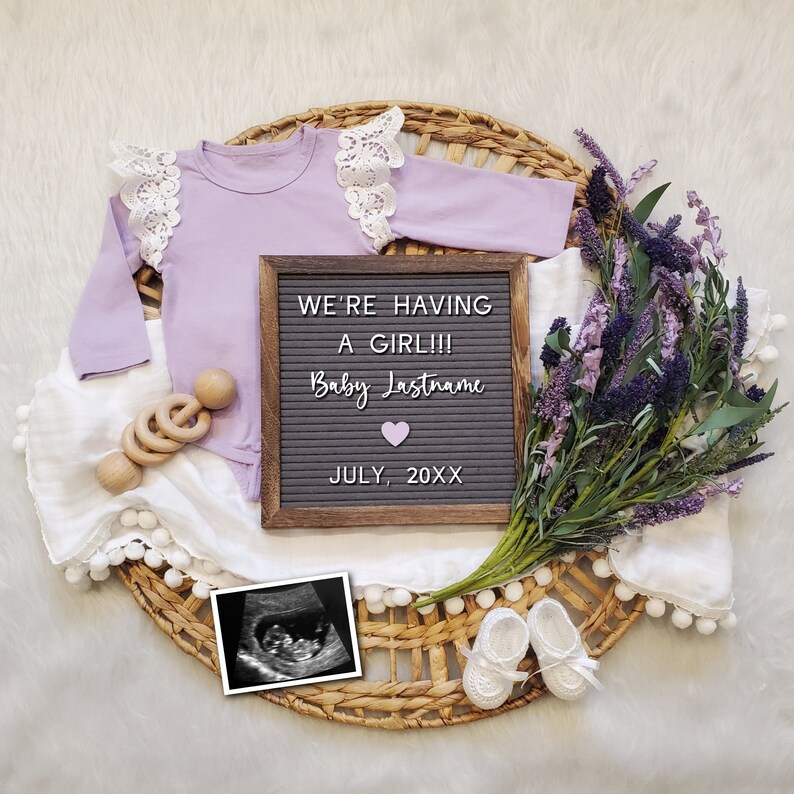 May include: A white blanket with a letter board that reads "We're having a girl!! Baby Lastname July, 20XX". A purple baby onesie with lace trim, a wooden rattle, a bouquet of lavender, and a black and white ultrasound image are also included.