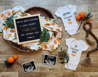 Op de afbeelding: Een zwart letterbord met de tekst "We've been keeping a juicy secret... 2 little cuties are joining our family!" Twee witte rompertjes met oranje fruitmotieven. Eén rompertje zegt "Arriving February 20XX" en het andere zegt "The Lastname Twins".