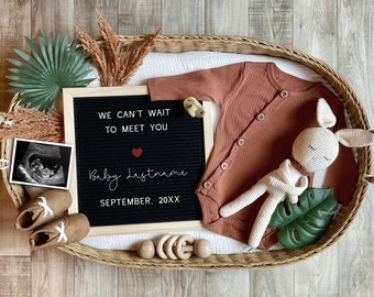 Op de afbeelding: Een zwart letterbord met de tekst "We can't wait to meet you Baby Lastname September, 20XX". Een rood hartje staat naast het woord "Baby". Het letterbord is omringd door een rieten mand met een bruine romper, een wit gehaakt konijn, een klein echografiebeeld en andere decoratieve items.