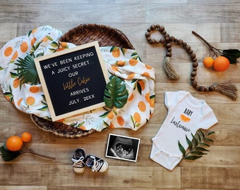 Op de afbeelding: Een zwangerschapsaankondiging met een letterbord waarop staat "We've been keeping a juicy secret. Our little cutie arrives July, 20XX". Er is een wit rompertje met de tekst "Baby Lastname" en een afbeelding van een sinaasappelschijf. Er is ook een baby-outfit, een echo-foto en een houten kralenketting met kwastjes.