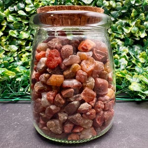 May include: A clear glass jar filled with a variety of colorful, polished stones. The stones are primarily shades of red, orange, and brown. The jar has a cork stopper and is set against a backdrop of green foliage.