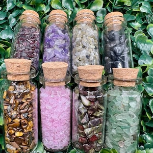 May include: Eight small glass bottles with cork stoppers filled with various types of colorful crystals and gemstones. The bottles are arranged in a row on a green leafy background.