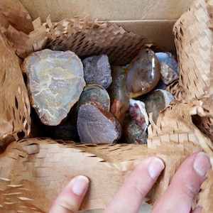 May include: A collection of various raw, unpolished stones and minerals in shades of brown, gray, and tan, nestled inside a cardboard box lined with brown honeycomb-patterned protective paper packaging for secure storage and transport.