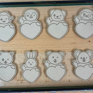 May include: Eight wooden cutouts of different animals, each holding a heart-shaped sign. The animals include a monkey, penguin, dog, sloth, cat, rabbit, bear, and koala. The cutouts are all in a light brown color and have a simple, cartoon-like style.