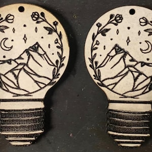 Op de afbeelding: Twee houten ornamenten in de vorm van een gloeilamp. Elk heeft een bergketen, een halve maan, sterren en bloemelementen in een cirkelvormig frame. De basis van elke lamp heeft een gestructureerd, donkerbruin ontwerp.