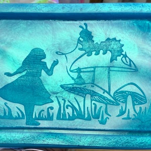 Könnte beinhalten: Ein blaugrünes Tablett mit einem erhabenen Design von Alice im Wunderland, die in einem Feld mit Pilzen steht. Das Design ist in einem helleren Blaugrünschatten gehalten.