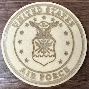 May include: A wooden coaster featuring the United States Air Force emblem. The emblem depicts a bald eagle with outstretched wings, a shield with a star and a cross, and the words "United States Air Force".