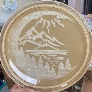 May include: A round, wooden serving tray with a clear glass insert. The tray features a laser-etched design of a mountain range, a lake, and a sun with stars.