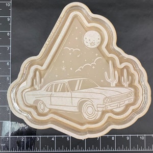 Può includere: Un ritaglio in legno bianco di un'auto d'epoca che guida attraverso un paesaggio desertico con una luna crescente e stelle. Il ritaglio è stratificato con un bordo bianco e il testo "THE ACRYLIC COTTAGE" è visibile nell'angolo in alto a sinistra.