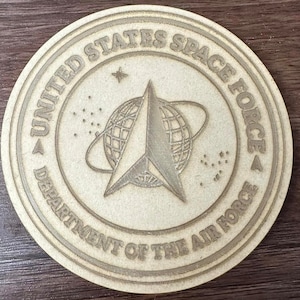 Puede incluir: Un posavasos redondo de madera con un diseño grabado en marrón del logotipo de la Fuerza Espacial de los Estados Unidos. El logotipo presenta una estrella con un globo terráqueo en su interior, rodeado de las palabras "United States Space Force" y "Department of the Air Force".