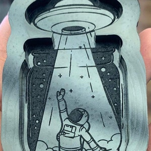 May include: A black and white illustration of an astronaut reaching up towards a UFO inside a mason jar. The UFO is emitting a beam of light. The astronaut is wearing a spacesuit and has a backpack. The background is a starry night sky.