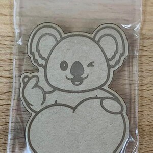 May include: A brown koala bear with a heart-shaped outline, holding a heart-shaped blank card. The koala is winking and has a happy expression.