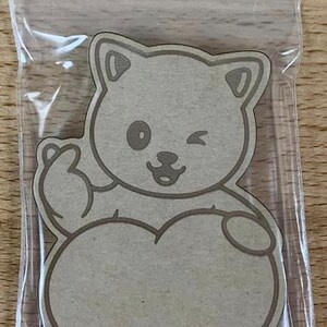 May include: A light brown cat-shaped wooden cutout winking and holding a heart. The cutout is inside a clear plastic bag with a zip closure. The cat has a simple outline and a heart-shaped body.