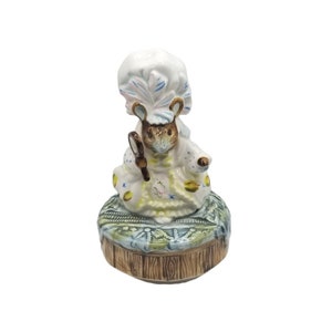 May include: A ceramic figurine of a mouse dressed in a white bonnet and gown, holding a small magnifying glass. The mouse is seated on a decorative base with a wooden-like appearance. The figurine has a vintage aesthetic.