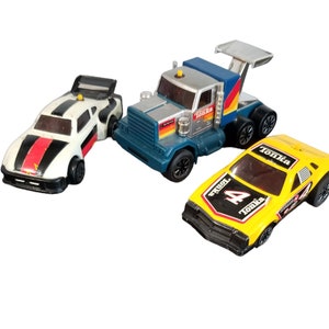 May include: Three vintage Tonka toy vehicles: a white race car with black and red stripes, a blue and silver semi-truck, and a yellow race car with the number 4. All toys have black wheels and are on a white background.