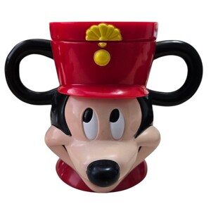 May include: A red and black Mickey Mouse-shaped mug with two black handles. The mug features Mickey's face, with a red hat and a yellow emblem. The mug is made of a glossy material and is designed to resemble the iconic Disney character.