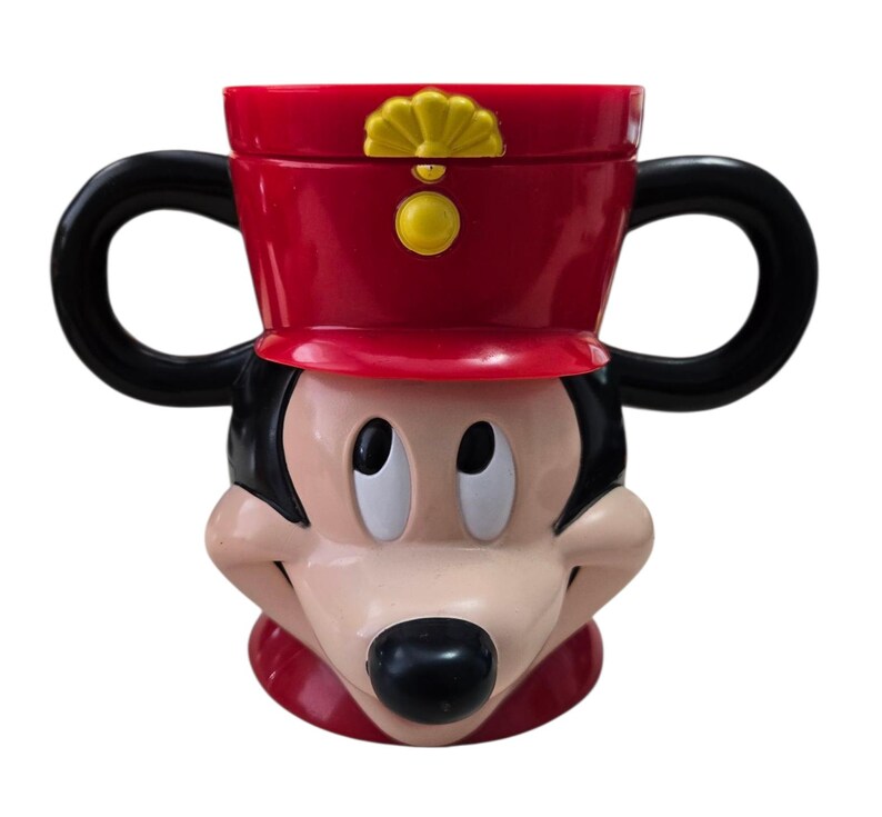 May include: A red and black Mickey Mouse-shaped mug with two black handles. The mug features Mickey's face, with a red hat and a yellow emblem. The mug is made of a glossy material and is designed to resemble the iconic Disney character.