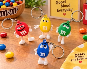以下が含まれることがあります： 赤、黄、緑、青のM&M'sキャラクターキーホルダー4点。それぞれ白い小文字のm、白い腕、白い靴が特徴です。キーホルダーには銀色の金属リングが付いています。木製の表面にはM&M'sのキャンディが散らばっています。
