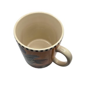 以下が含まれることがあります： ベージュの内側とハンドルが付いたセラミックコーヒーマグ。外側は茶色のデザインで、「NEWS」の文字が見えます。縁には白と黒の市松模様があります。