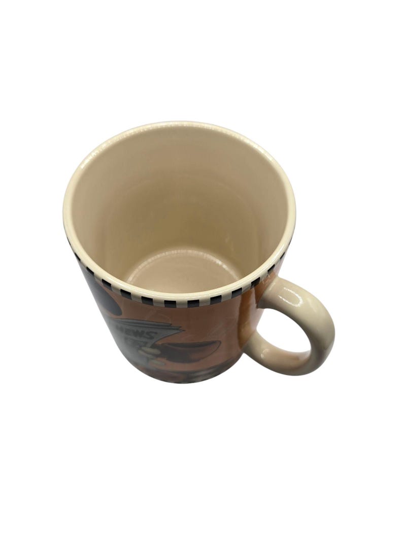 以下が含まれることがあります： ベージュの内側とハンドルが付いたセラミックコーヒーマグ。外側は茶色のデザインで、「NEWS」の文字が見えます。縁には白と黒の市松模様があります。