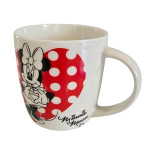 Op de afbeelding: Witte keramische mok met Minnie Mouse. De mok heeft een rood hartvormig ontwerp met witte stippen. Minnie Mouse is afgebeeld in zwart, wit en rood. De mok heeft een gebogen handvat en de woorden "Minnie Mouse" zijn in cursief geschreven.
