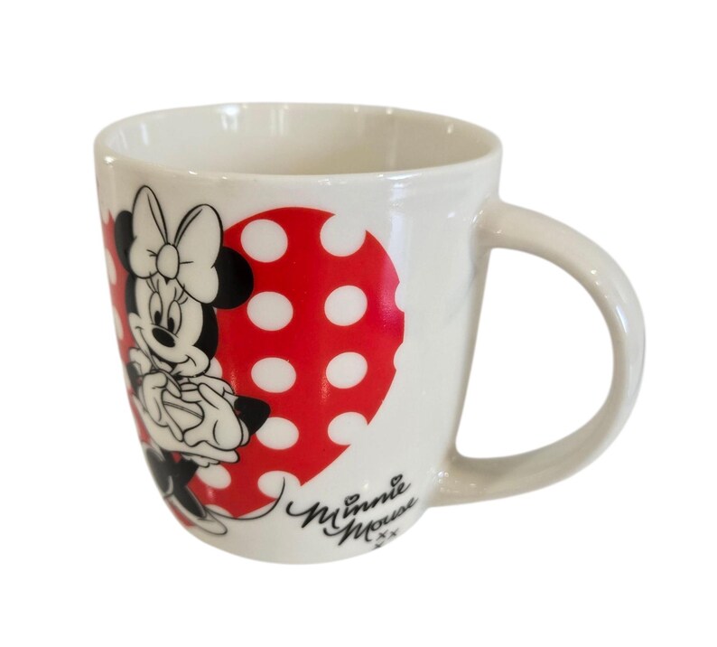 Op de afbeelding: Witte keramische mok met Minnie Mouse. De mok heeft een rood hartvormig ontwerp met witte stippen. Minnie Mouse is afgebeeld in zwart, wit en rood. De mok heeft een gebogen handvat en de woorden "Minnie Mouse" zijn in cursief geschreven.