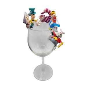 May include: A clear wine glass decorated with characters from Alice in Wonderland. The Mad Hatter, Cheshire Cat, Alice, and the White Rabbit are positioned around the rim. The figures are colourful and detailed, creating a whimsical design.