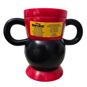 May include: A red, black, and white Disney "On Ice" mug with two black handles. The mug has a rounded base and a cylindrical top. A yellow sticker with text is visible on the side.