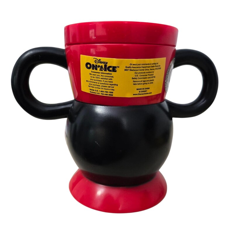 May include: A red, black, and white Disney "On Ice" mug with two black handles. The mug has a rounded base and a cylindrical top. A yellow sticker with text is visible on the side.