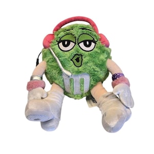 May include: A green M&M's soft toy wearing pink headphones, silver boots, and wrist cuffs. The character has a silver microphone and the letter "M" on its front. The plush toy has a kissy face.