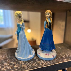 Puede incluir: Dos figuras que representan personajes con cabello rubio. Una figura viste un vestido largo de color azul claro con una capa. La otra tiene el cabello trenzado y viste un corpiño negro con una falda larga azul con pequeños detalles florales.