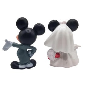 May include: Two figurines of Mickey and Minnie Mouse in wedding attire. Mickey is in a green suit with white gloves, gesturing with his hand. Minnie is in a white wedding dress and veil, holding a bouquet of red roses.