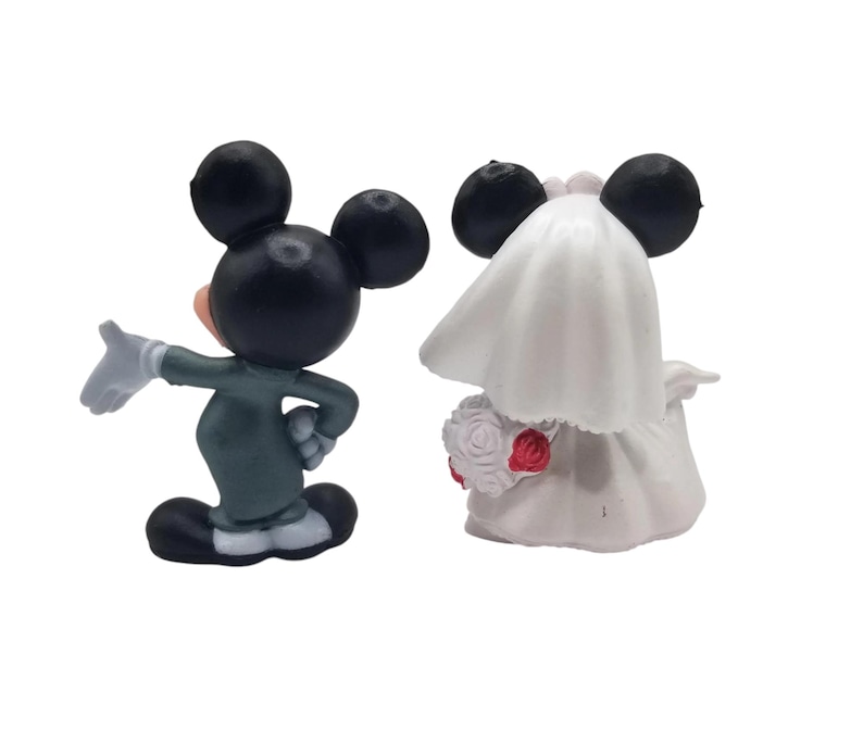 May include: Two figurines of Mickey and Minnie Mouse in wedding attire. Mickey is in a green suit with white gloves, gesturing with his hand. Minnie is in a white wedding dress and veil, holding a bouquet of red roses.