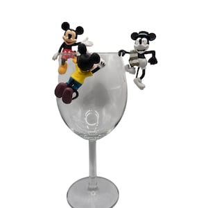 May include: A clear wine glass with three Mickey Mouse figurines perched on the rim. One figurine wears a yellow shirt, another a red shirt, and the third a scarf. The figurines are black, white, and red.