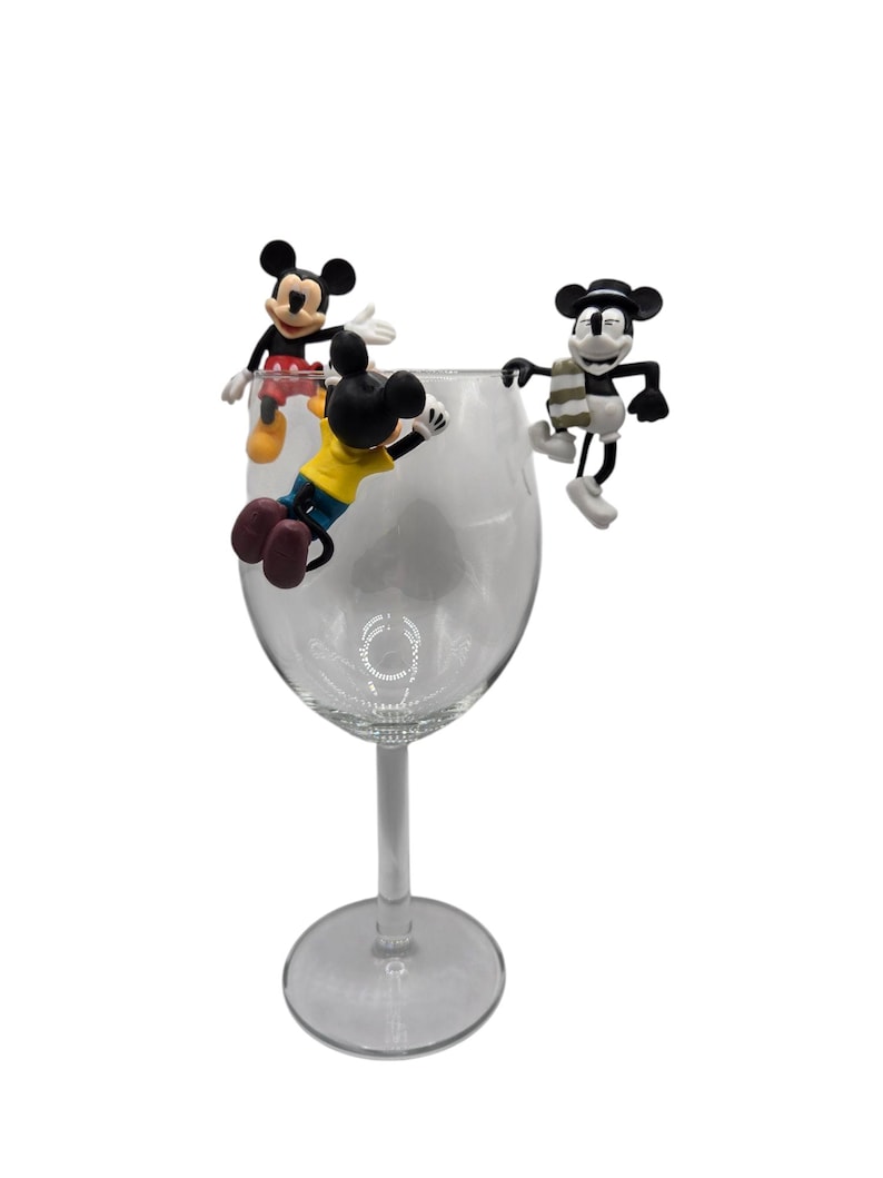 May include: A clear wine glass with three Mickey Mouse figurines perched on the rim. One figurine wears a yellow shirt, another a red shirt, and the third a scarf. The figurines are black, white, and red.