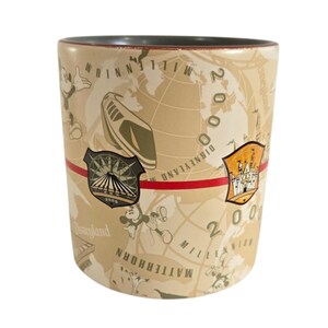 Pode incluir: Caneca cil&iacute;ndrica de cer&acirc;mica com fundo bege com um design de mapa-m&uacute;ndi e o texto "2000 Millennium Disneyland". A caneca tem uma faixa vermelha e imagens do Mickey Mouse e atra&ccedil;&otilde;es da Disney.