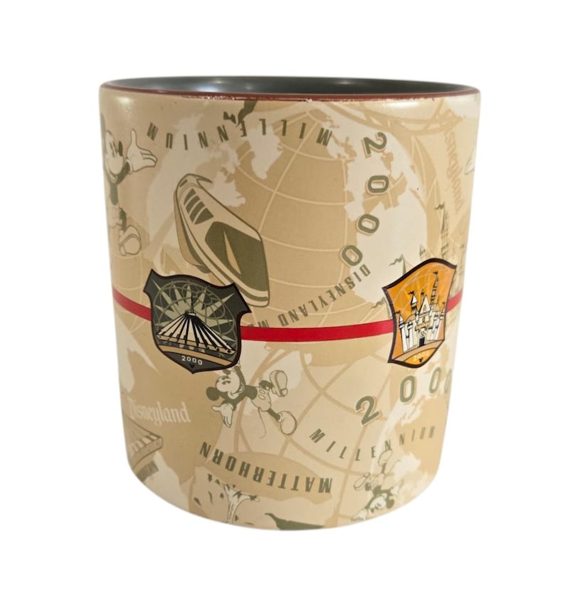 Pode incluir: Caneca cil&iacute;ndrica de cer&acirc;mica com fundo bege com um design de mapa-m&uacute;ndi e o texto "2000 Millennium Disneyland". A caneca tem uma faixa vermelha e imagens do Mickey Mouse e atra&ccedil;&otilde;es da Disney.