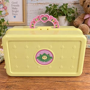 May include: A pale yellow plastic carrying case featuring a grid pattern with small embossed apple shapes. The case has a pink heart-shaped handle with floral accents and a central circular decal depicting a cabbage-like character with a face.