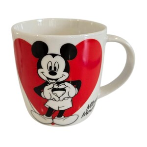 Op de afbeelding: Witte keramische mok met Mickey Mouse in een rood hart. Mickey is afgebeeld in zwart, wit en rood, met zijn handen in een hartvorm. De mok heeft een gebogen oor en de woorden "Minnie Mouse" staan op de zijkant.