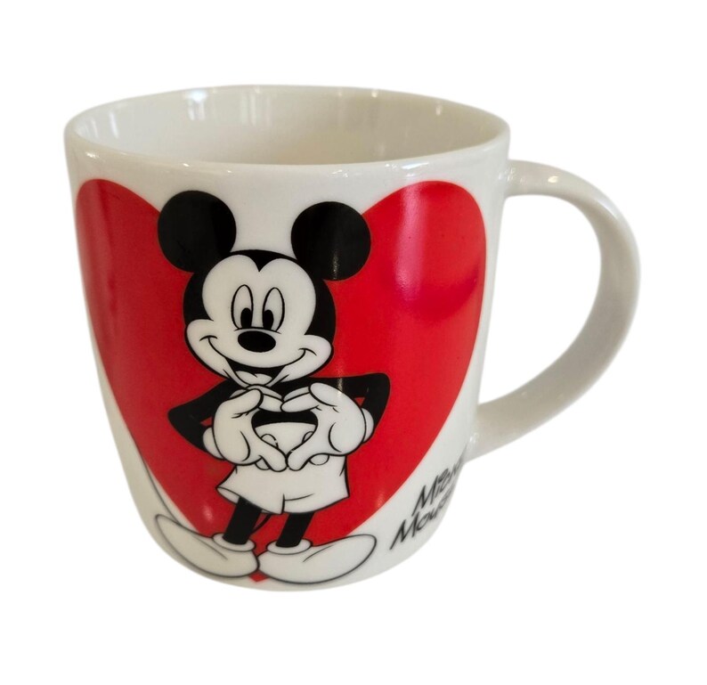 Op de afbeelding: Witte keramische mok met Mickey Mouse in een rood hart. Mickey is afgebeeld in zwart, wit en rood, met zijn handen in een hartvorm. De mok heeft een gebogen oor en de woorden "Minnie Mouse" staan op de zijkant.