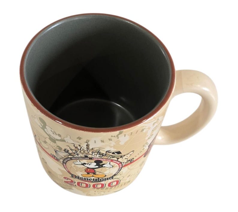 Pode incluir: Caneca de cer&acirc;mica creme com um design de mapa e as palavras "Disneyland 2000" com Mickey Mouse. A caneca tem uma al&ccedil;a bege e um interior cinza escuro. A borda &eacute; marrom.