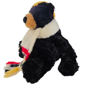 May include: A plush black bear with a tan muzzle and yellow accents on its head. The bear wears a cream-colored scarf with red, blue, and yellow stripes. The bear is sitting down.