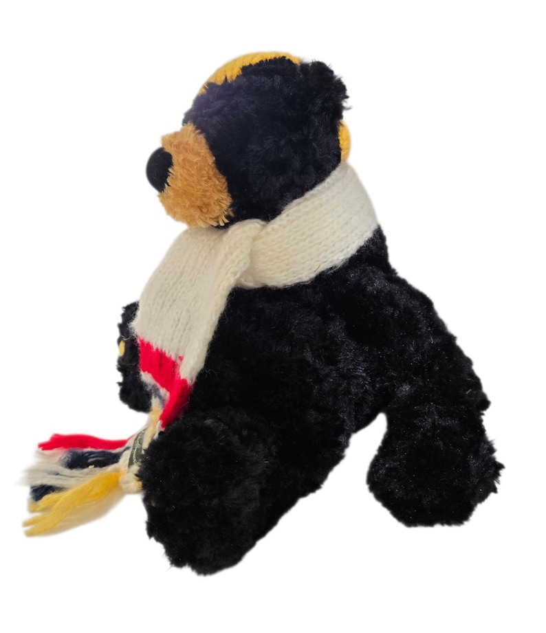 May include: A plush black bear with a tan muzzle and yellow accents on its head. The bear wears a cream-colored scarf with red, blue, and yellow stripes. The bear is sitting down.