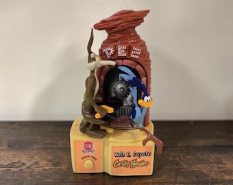 Può includere: Un distributore di caramelle in plastica Wile E. Coyote Candy Hander con una figura di coyote marrone e una figura di Beep Beep blu davanti a una formazione rocciosa rossa con il testo PEZ. La base è gialla con un pulsante e il testo Wile E. Coyote Candy Hander.